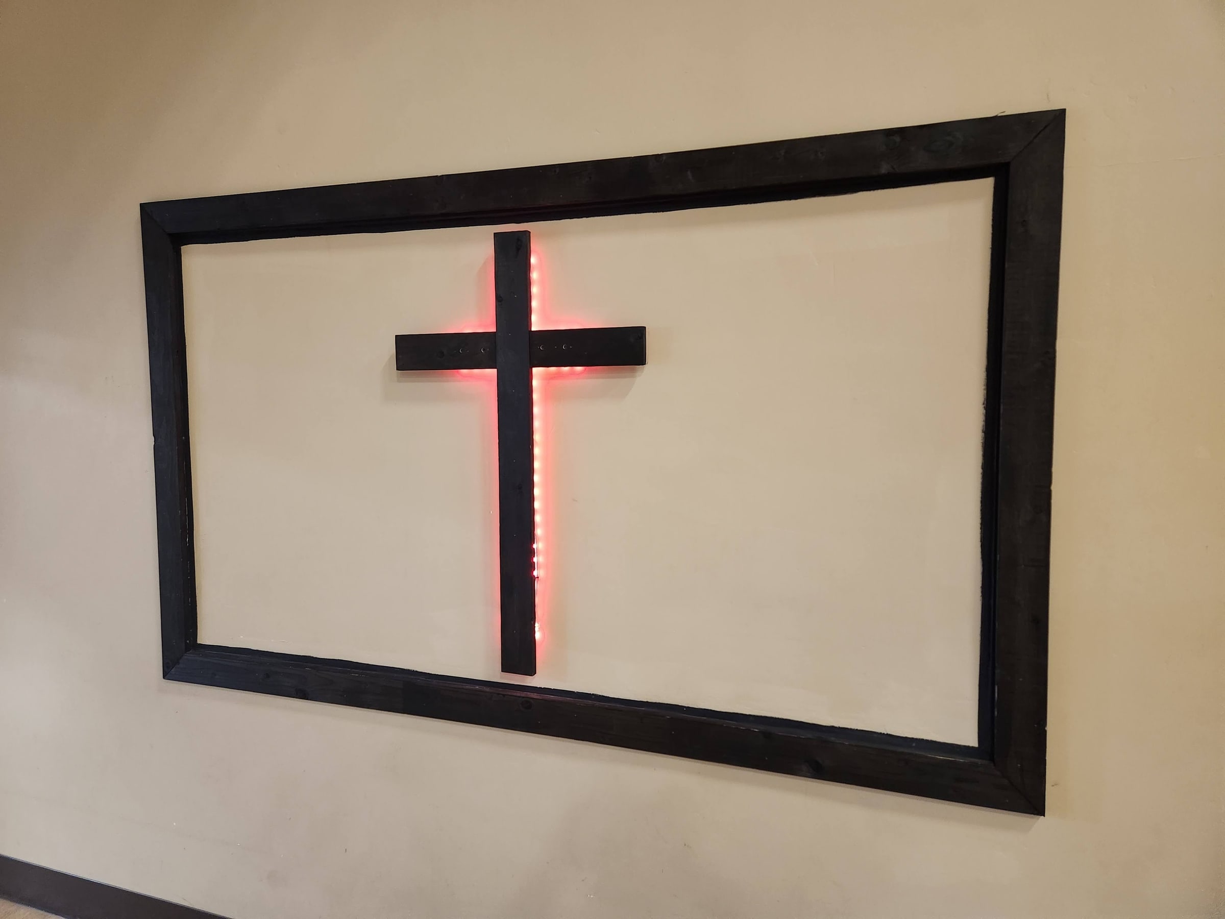 A wooden cross lit from behind, mounted in a dark wooden frame on a white wall