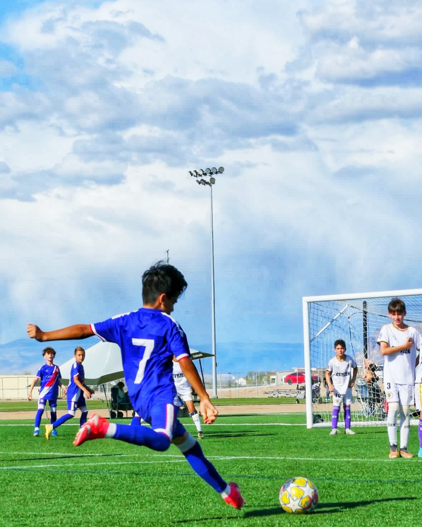 A young player in a blue number 7 jersey kicks the ball toward the goal on an outdoor field