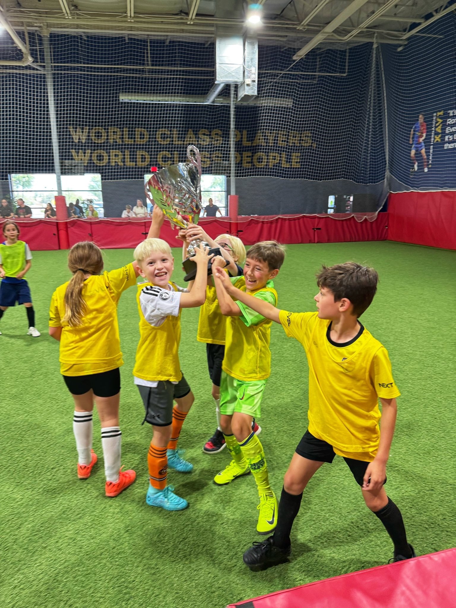 Young players in yellow jerseys cheer and lift a trophy on the indoor turf field