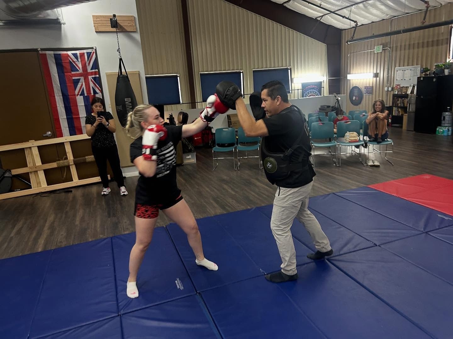 Two adults practice boxing drills with pads on the blue mats at On The Rock Martial Arts