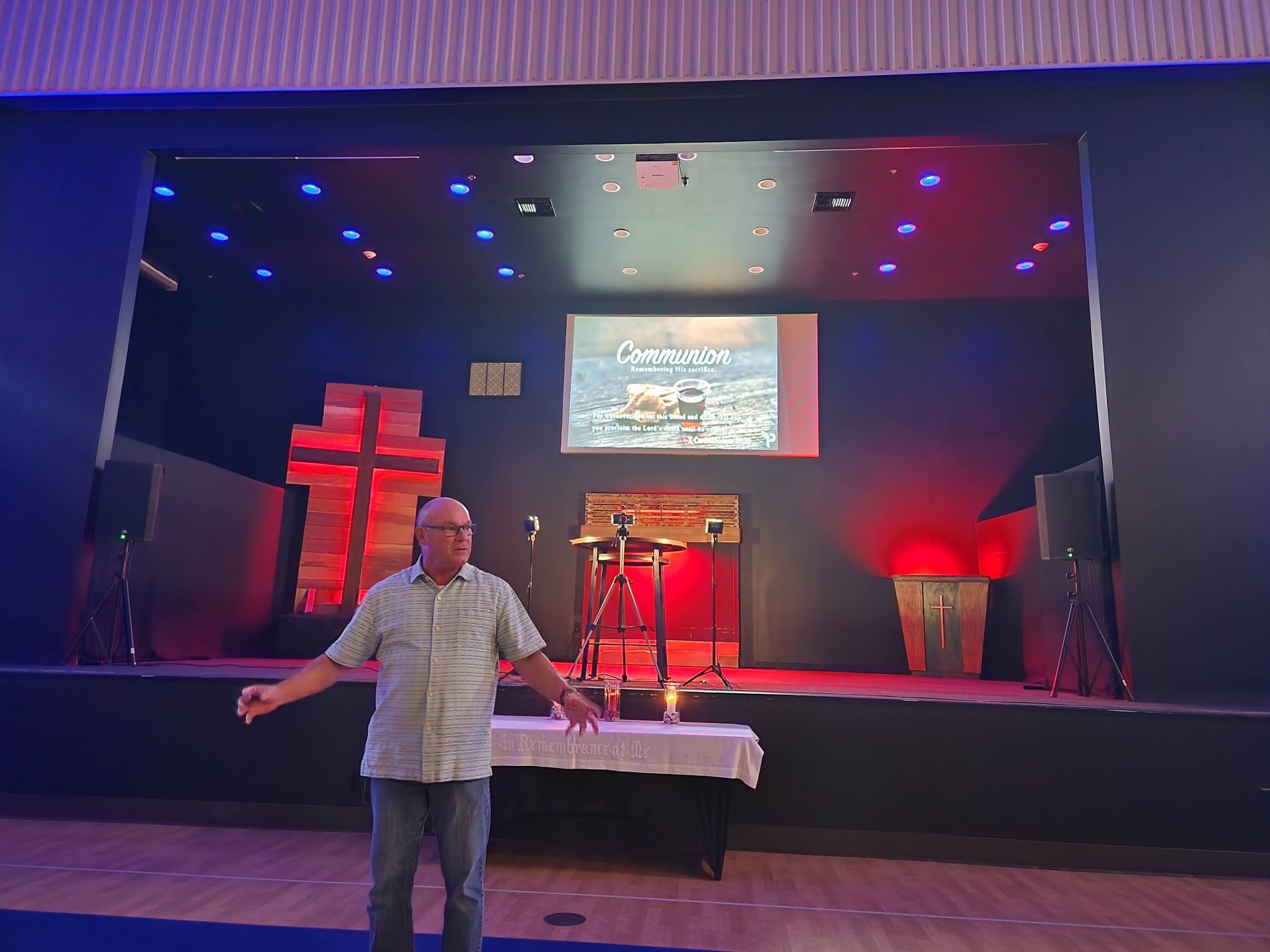 Pastor Jim speaks on stage beside a wooden cross with “Communion” on the screen behind him