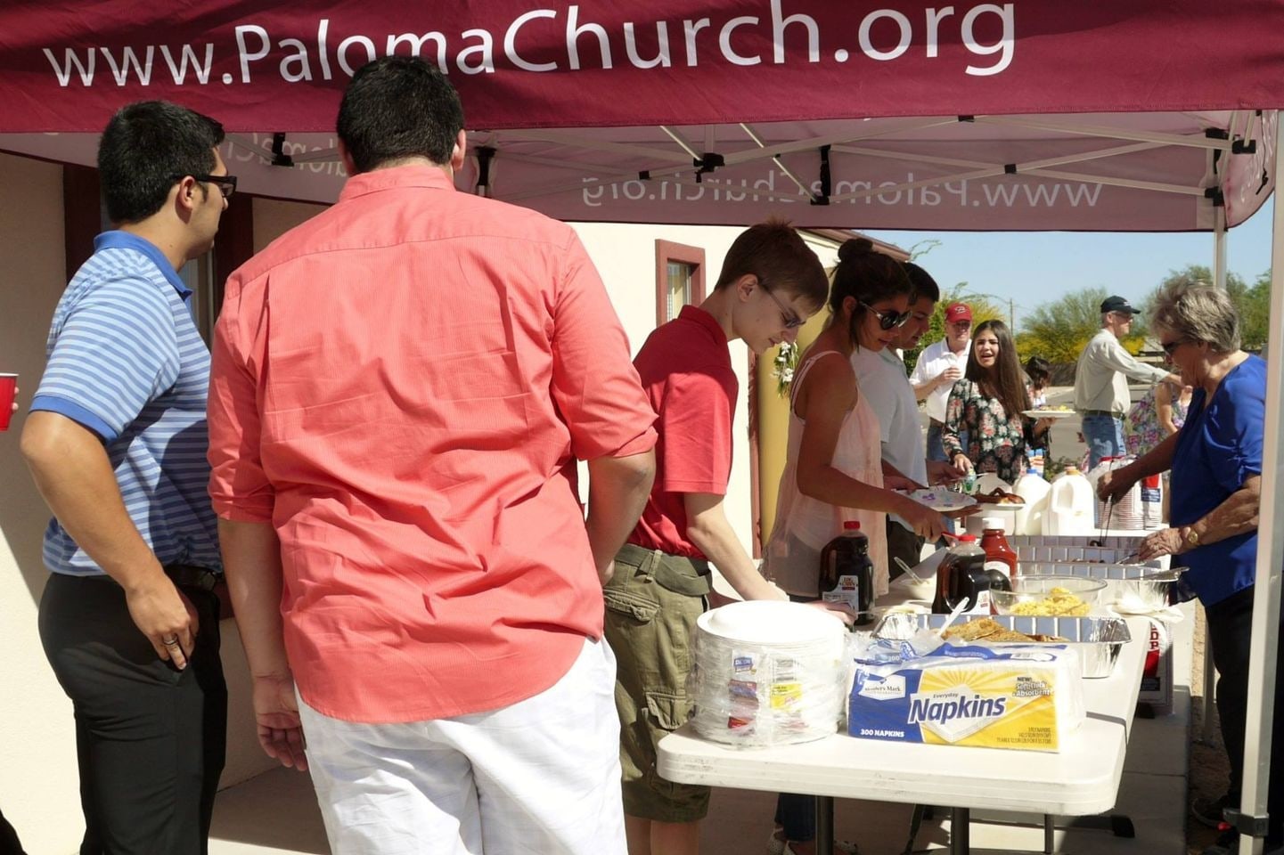 Volunteers serve food and drinks from tables at an outdoor Paloma Church event
