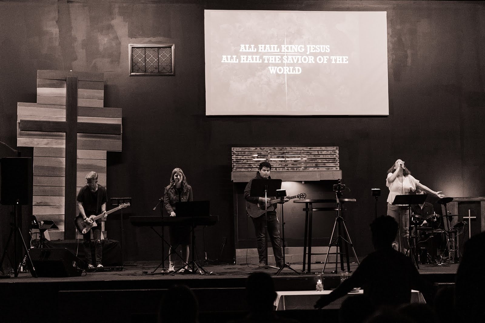 The worship band plays on stage beneath a wooden cross while the screen reads “All hail King Jesus”