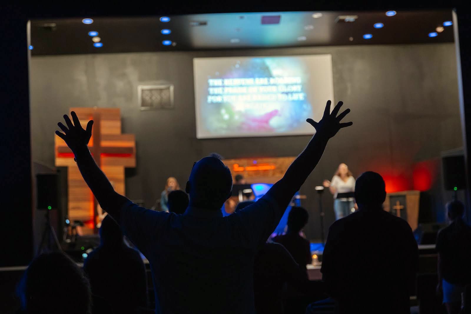 A person raises both arms in worship, silhouetted against the stage, cross and screen during a Sunday service