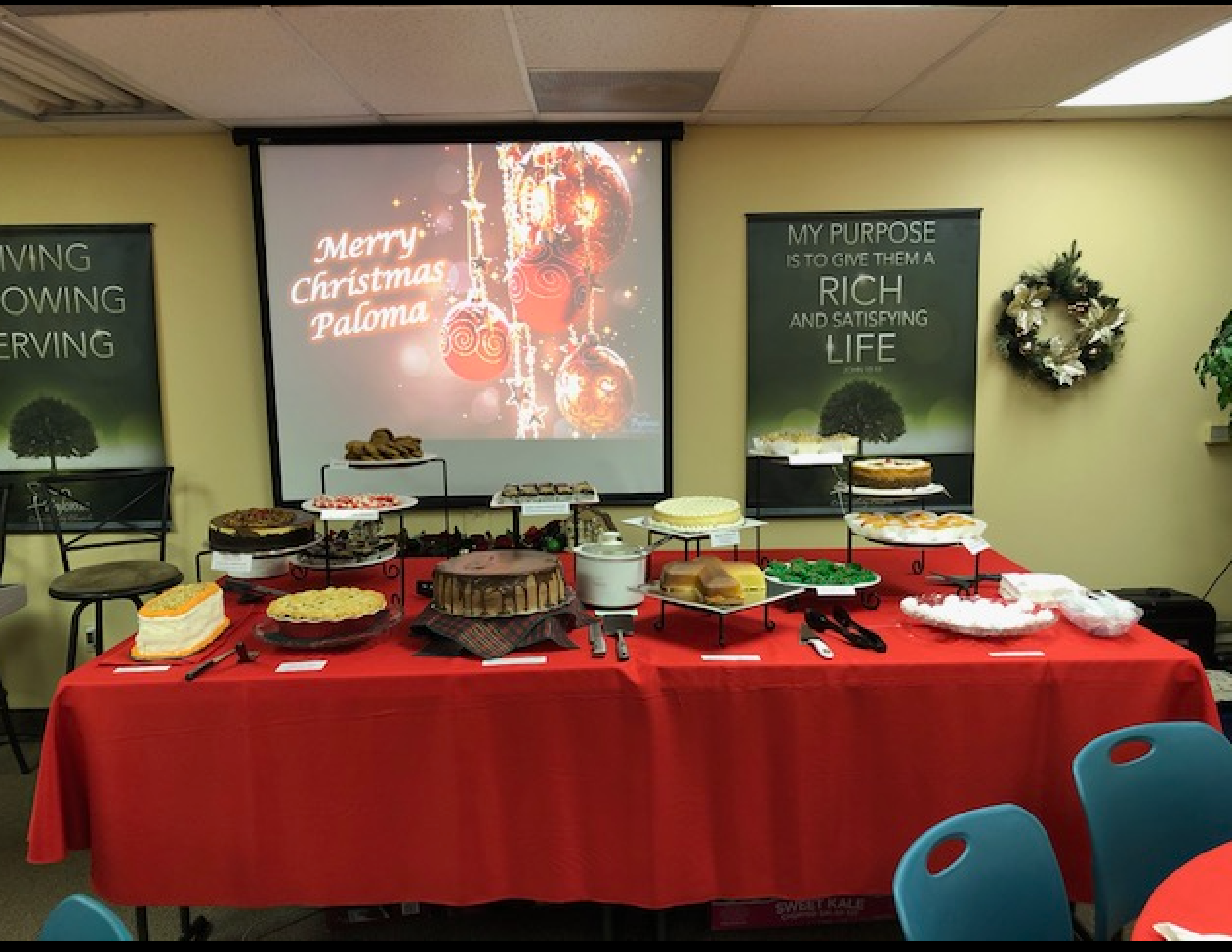 Paloma Table: a long table with a red cloth set with cakes, pies and cookies, in front of a screen reading “Merry Christmas Paloma”.